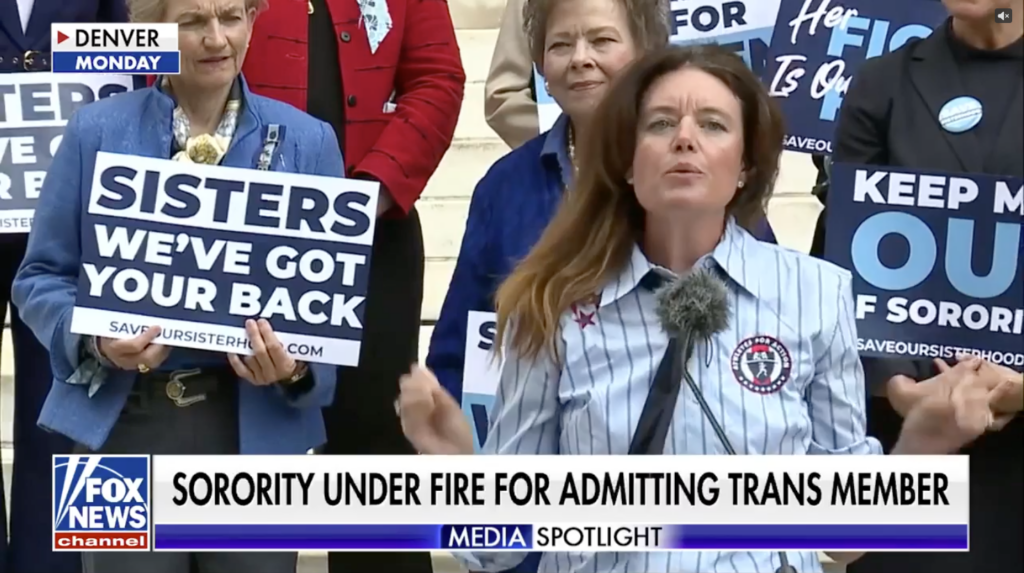 Speaker at a mic flanked by supporters holding blue signs reading 'SISTERS WE'VE GOT YOUR BACK' at a press event.